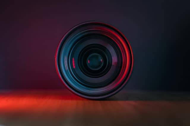Camera lens standing on a table, its rim lit with a red glow against a dark background