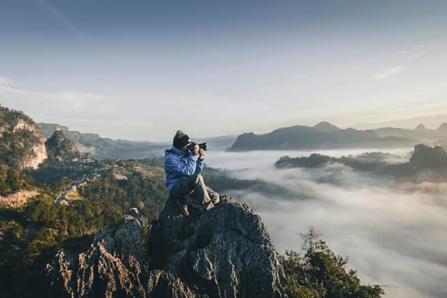 Photographer crouched on a rocky summit, shooting a valley filled with morning cloud