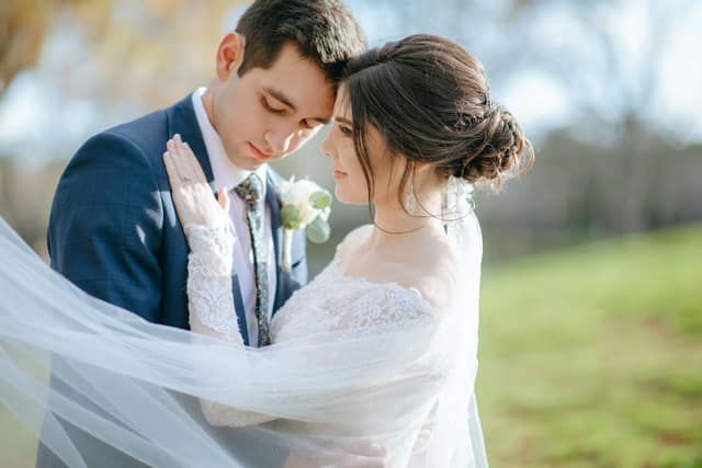 Bride and groom touching foreheads, wrapped in a flowing veil outdoors